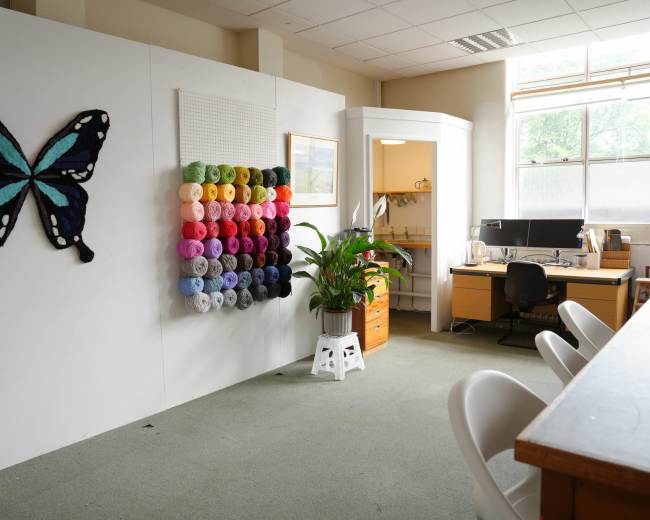 The image shows a craft room featuring a wall with colorful yarn organized in a grid pattern, a desk with a computer, and a large butterfly artwork on the wall.