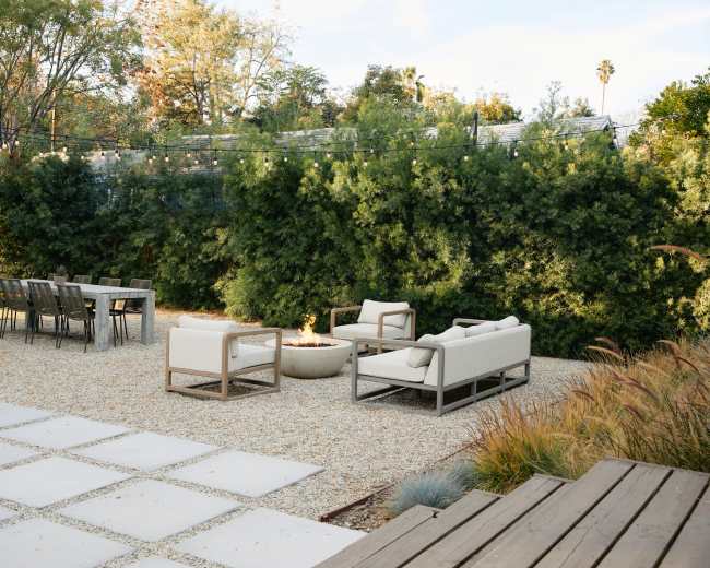 The image shows a landscaped outdoor space featuring a gravel area with a seating arrangement around a fire pit and a dining table nearby.