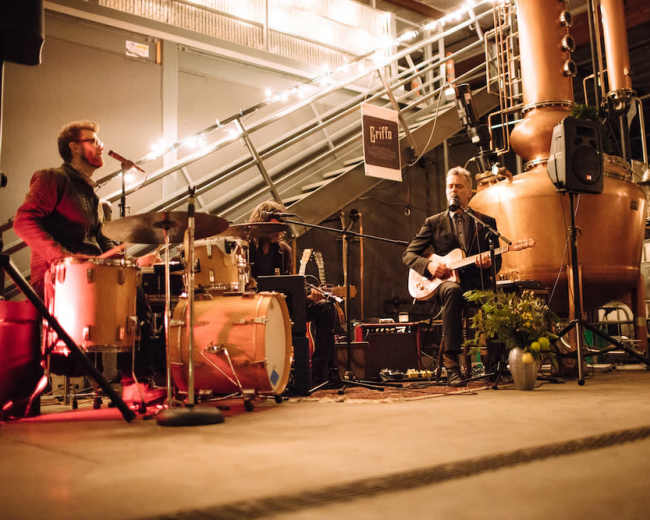 A band performs on a stage set up in a brewery, surrounded by brewing equipment and string lights.