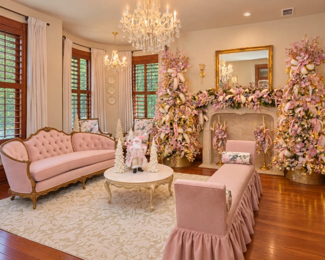 The room features elegant pink furniture and decor, with a centerpiece of decorated Christmas trees flanking a beautifully adorned fireplace.