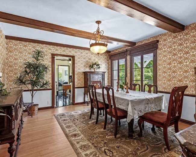 The image shows a dining room with a long table set for meal, surrounded by wooden chairs, against a backdrop of floral wallpaper and wooden beams on the ceiling.