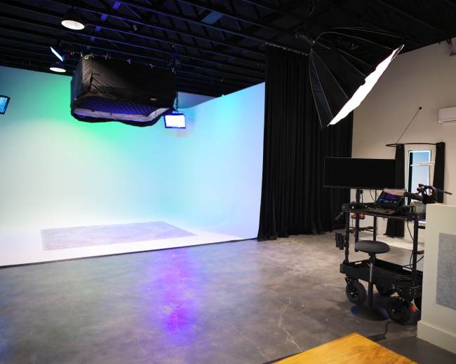 The image shows a professional photography or video studio with a seamless backdrop, lighting equipment, and a mobile workstation.