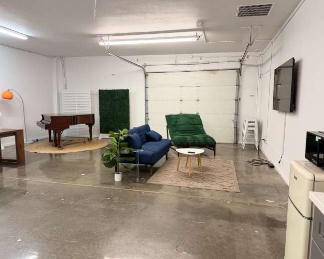 The image shows a spacious, minimalist room with polished concrete floors, featuring a blue sofa, a green lounge chair, a piano, a small table, and a plant, along with garage doors at one end.