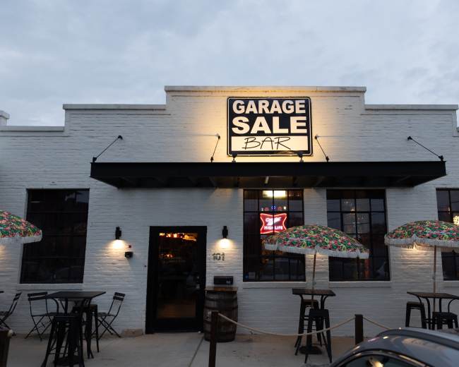 The image shows the exterior of a bar labeled "Garage Sale Bar" with outdoor seating and colorful umbrellas.