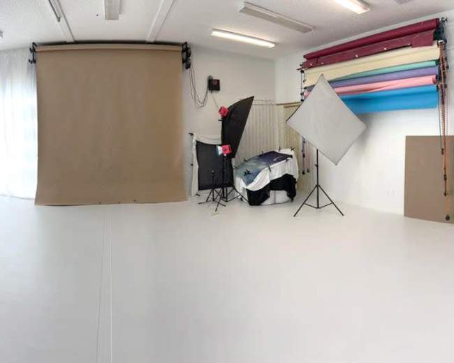 The image shows a spacious photo studio with white walls, a wooden door, and various backdrops in pastel colors.