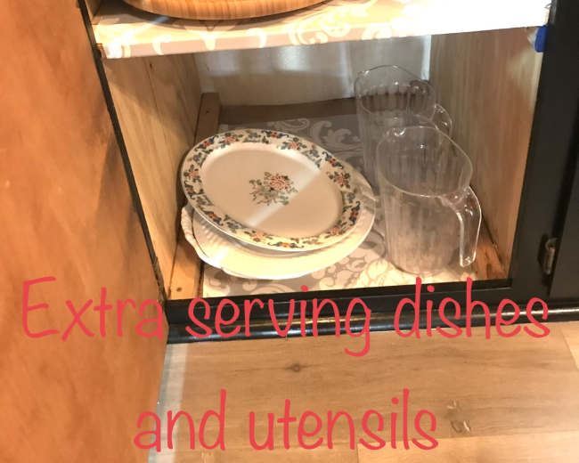 A wooden cabinet shelf holds a variety of serving dishes, utensils, and glass pitchers.