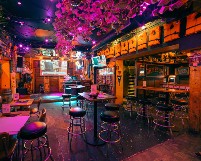 The image shows a vibrant bar interior with purple lighting, adorned with plants, displaying a projector screen at the back and a mix of high tables and stools.