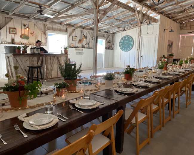 A long wooden table is set for a meal in a rustic indoor space featuring a bar, potted plants, and a large clock on the wall.