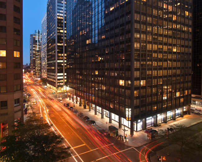 The image shows a view of tall glass buildings at dusk, with traffic lights creating streaks of light on the busy street below.