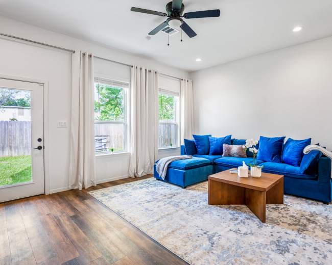 A bright living room features a blue sectional sofa, a wooden coffee table, and large windows with sheer curtains.