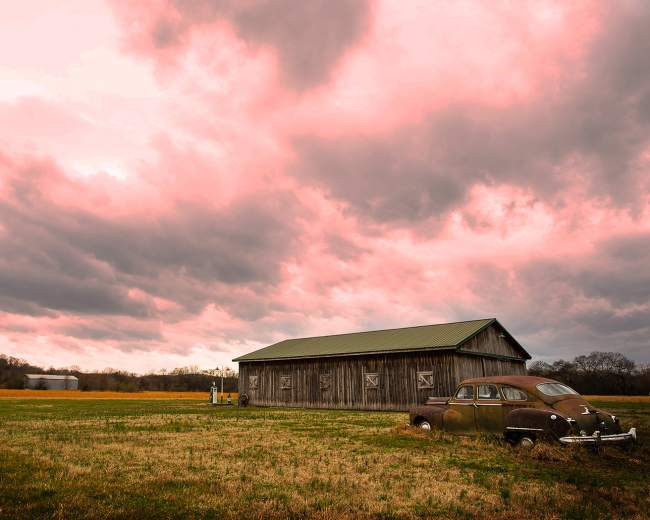 A weathered barn stands beside an old car in a grassy field under a dramatic pink and gray sky.