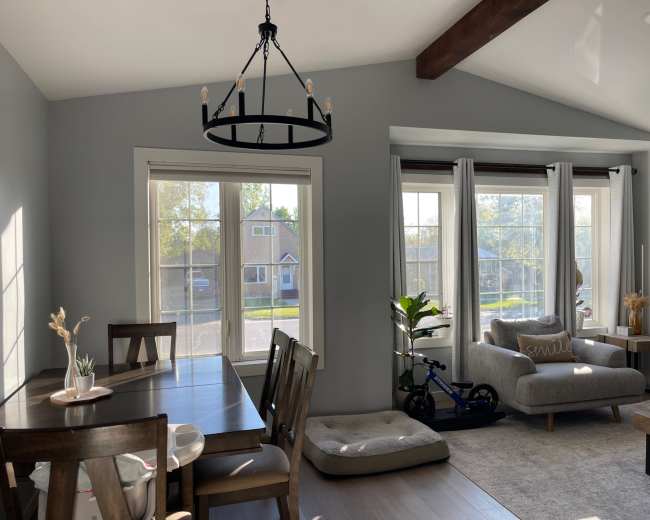 A brightly lit living space features a dining area with a wooden table, a cozy seating area with a gray sofa, and large windows showcasing a view of the outdoors.
