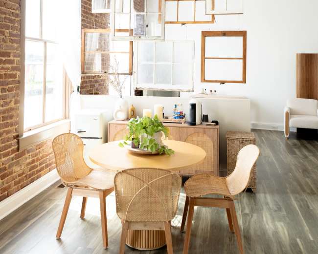 A round wooden dining table with four woven chairs is positioned in a bright room featuring exposed brick walls and large windows.