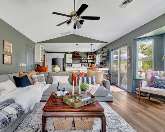 A cozy living room features a large gray sectional sofa, a wooden coffee table, and open access to a bright kitchen area.