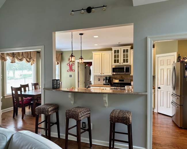 The image shows a kitchen area with a bar counter and three stools, connecting to an adjacent dining space and living room.