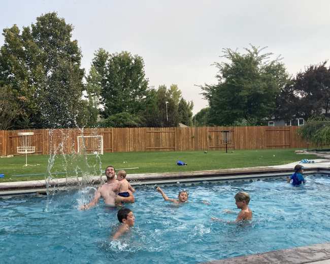 A group of children and an adult are playing in a swimming pool, with water splashing around as they enjoy the warm day.