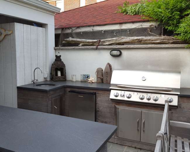 The image shows an outdoor kitchen area with a grill, sink, and countertop, surrounded by wooden walls and decorative elements.