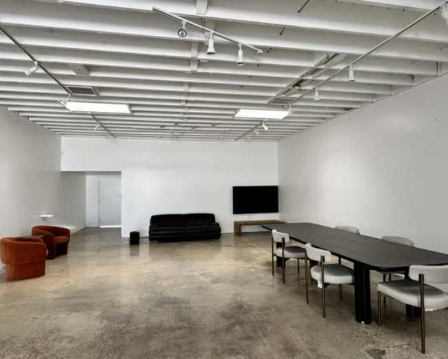 The image shows a spacious, minimalistic room with a black sofa, two orange chairs, a long table with white chairs, and a television against a white wall.