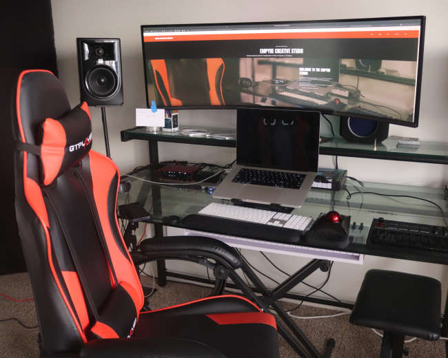 The image shows a modern workspace with an ergonomic gaming chair, a large curved monitor displaying a website, and various computer accessories on a glass desk.