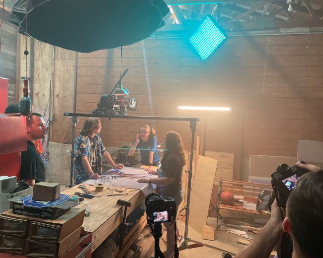 A film crew sets up a scene in a wood-paneled studio, with lighting equipment illuminating three people discussing on a table while cameras capture the action.