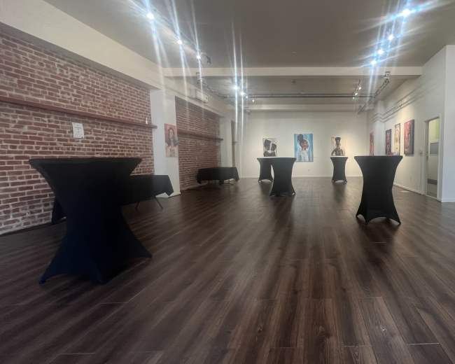 The image shows an art gallery with black cocktail tables set up on a polished wooden floor, surrounded by various paintings hanging on the white walls.