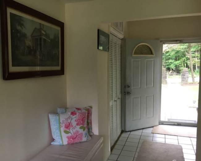 The image shows a light-colored entryway with a bench, a framed picture on the wall, and a door leading outside.