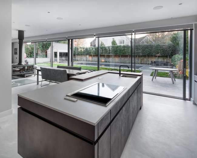 The image shows a modern kitchen with a large island, featuring a cooktop, overlooking a spacious outdoor area through expansive glass windows.
