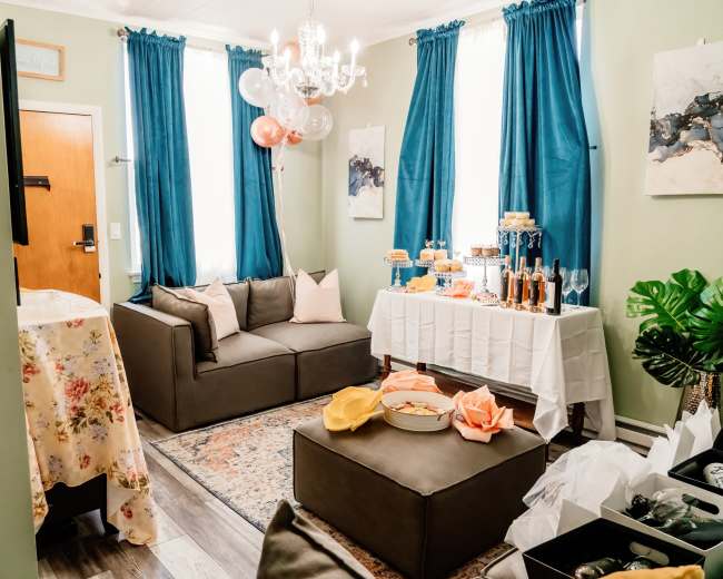 The living room features a grey sectional sofa, a decorative table with a white tablecloth holding dessert platters, and walls adorned with blue curtains and artwork.