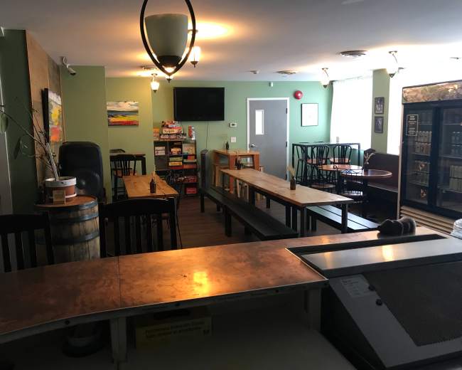 A spacious, empty café with wooden tables, chairs, and a counter, featuring shelves of beverages and games in the background.