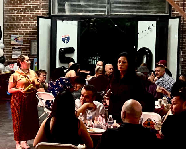 A group of people is seated at tables during a gathering in an industrial-style venue with exposed brick walls and balloons in the background.