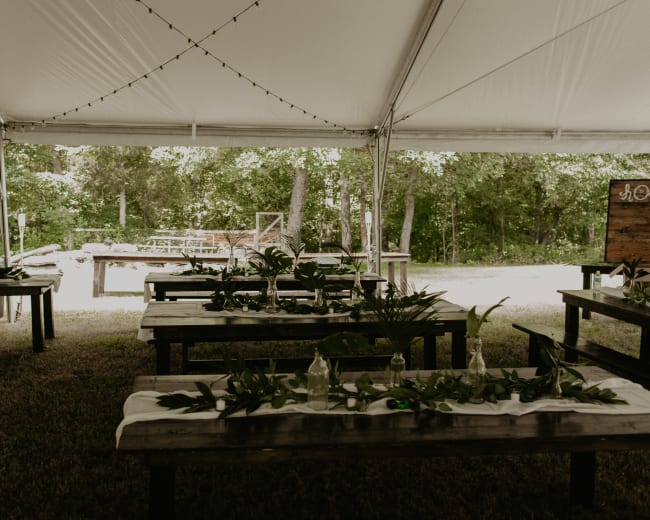 A banquet tent with wooden tables covered in white cloth and adorned with greenery is set up in a shaded outdoor area.