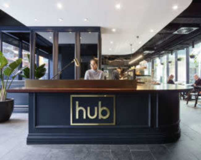 A sleek reception desk with the word "hub" prominently displayed, staffed by a person, in a modern, well-lit interior with seating areas in the background.
