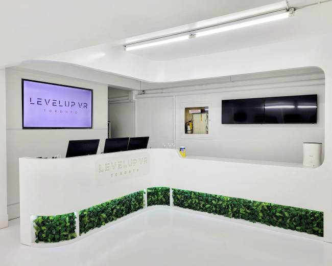 The image shows a modern reception area for Level Up VR in Toronto, featuring a sleek white counter with greenery at the base and a large screen displaying the company name.