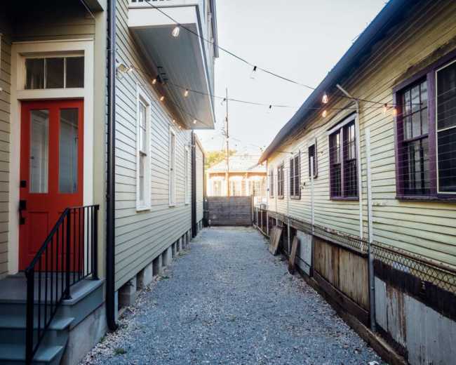 A narrow gravel pathway runs between two houses, one with a bright red door and the other with yellow siding, illuminated by string lights above.