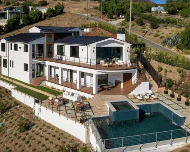 The image shows a modern two-story house situated on a hillside, featuring a swimming pool and landscaped outdoor area.