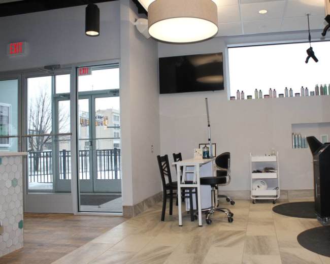 The image shows a modern salon interior with a reception area, a treatment table, and products displayed on shelves near large windows.