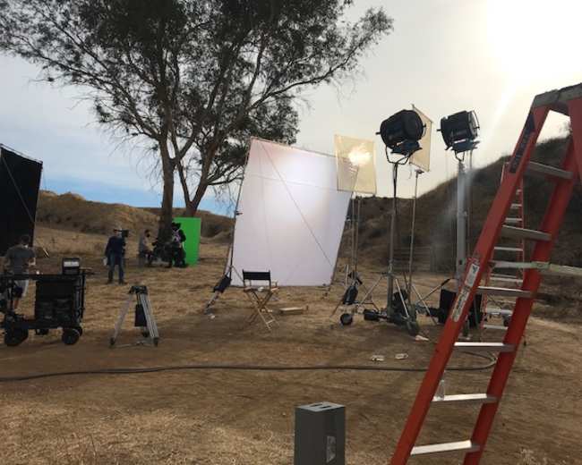 A film set is arranged outdoors with lighting equipment, a green screen, and a ladder positioned next to a tree on a dry hillside.