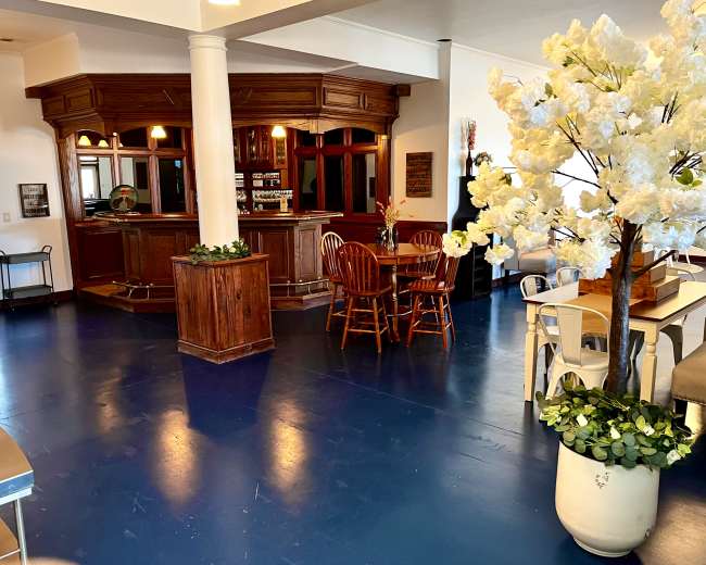 The image shows a spacious interior with a bar area, wooden seating, and a decorative flower arrangement.