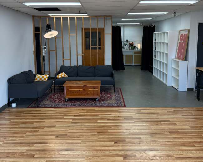 The image shows a spacious room featuring a seating area with a sofa and coffee table, wood flooring, and an open kitchen area in the background.
