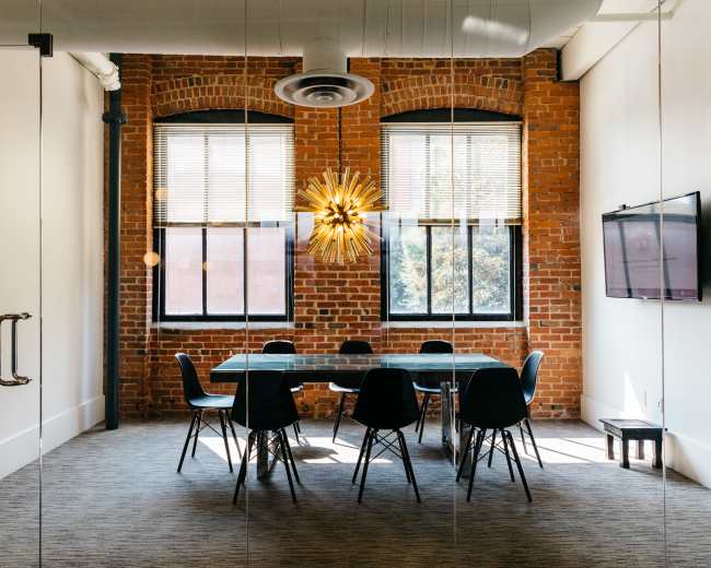 A modern conference room features a large table surrounded by black chairs, with exposed brick walls and large windows allowing natural light to enter.