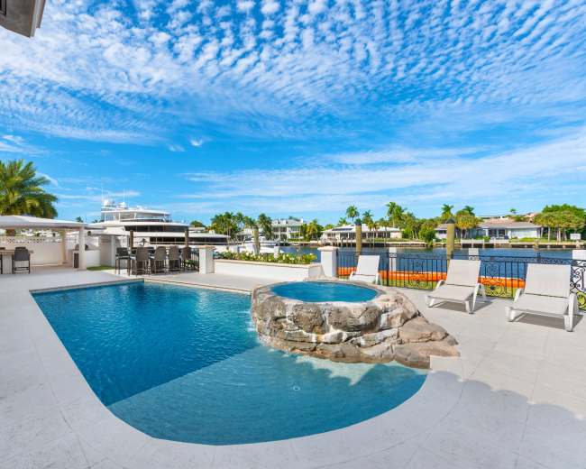 A backyard features a swimming pool with a rock waterfall and a spa overlooking a canal, surrounded by palm trees and a patio area.