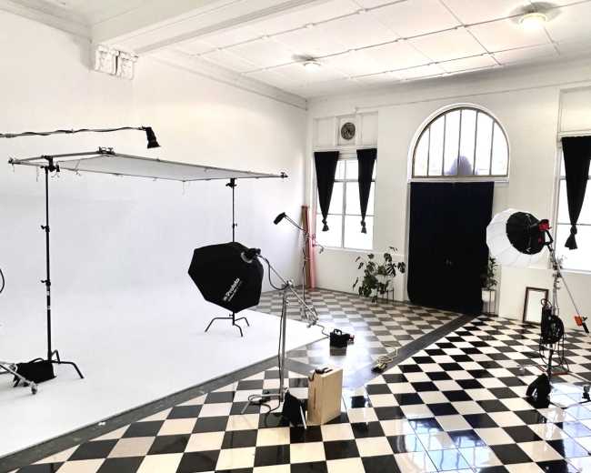 The image shows a photography studio with a white backdrop, lighting equipment, and a checkered floor.