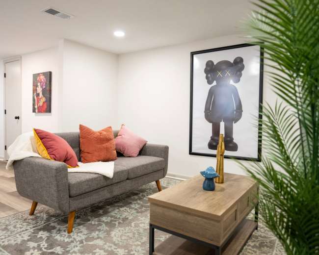 The image shows a modern living space with a gray sofa adorned with colorful cushions, a wooden coffee table, and a large framed artwork on the wall.