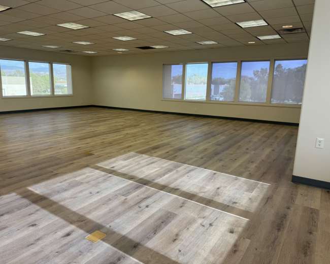The image shows a spacious empty office room with wooden flooring and several large windows allowing natural light to enter.