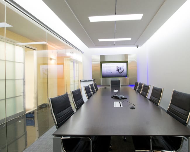 A modern conference room features a long table surrounded by black chairs, with a wall-mounted screen displaying information.