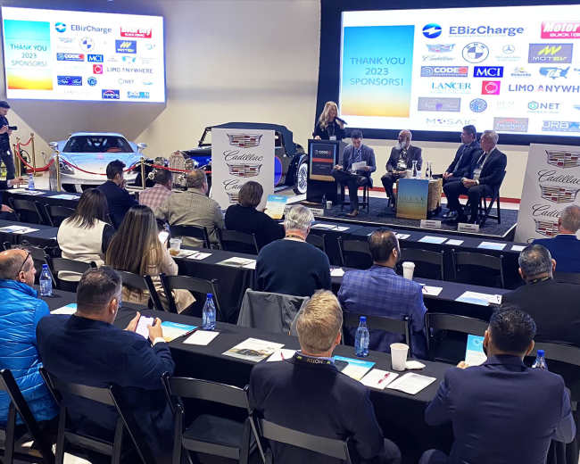 A panel discussion is taking place in front of an audience at an event featuring several classic cars and promotional screens in the background.