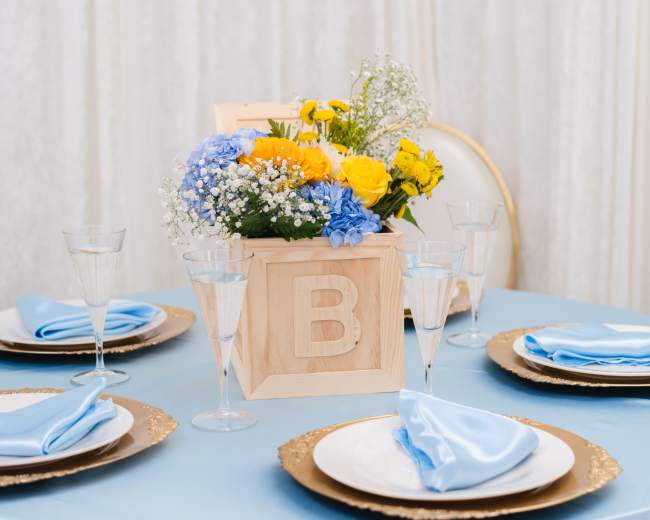 A table is set with blue tablecloth, decorative plates, and a wooden box of flowers in the center.