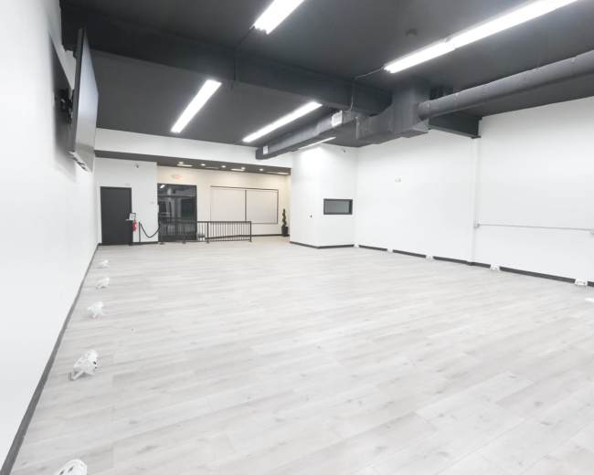 The image shows a large, empty room with light-colored wooden flooring and white walls, featuring a wall-mounted TV and a hand sanitizer station.
