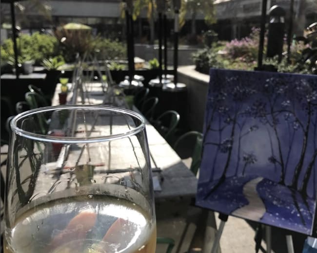 A glass of beverage in the foreground with a painted canvas of a snowy path and trees in the background, set in an outdoor dining area.