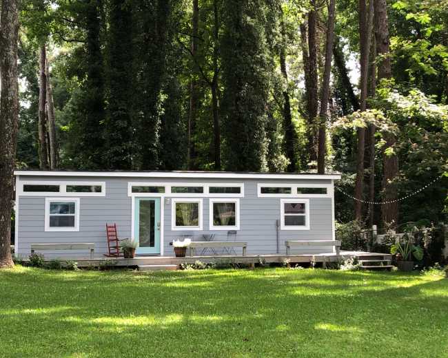 A modern, single-story house with a sloped roof is surrounded by tall trees and sits on a grassy lawn.